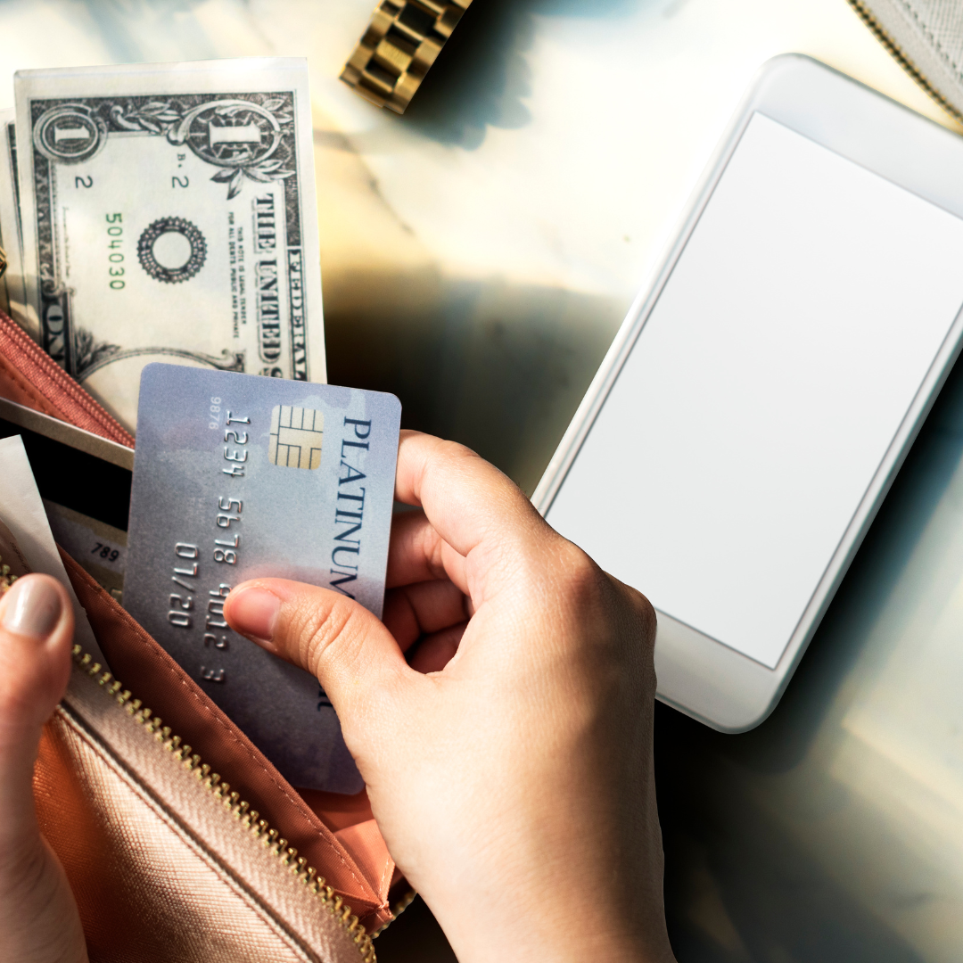 View of hands holding an open wallet with cash and a credit card. A smartphone sits on the table beside the hand.