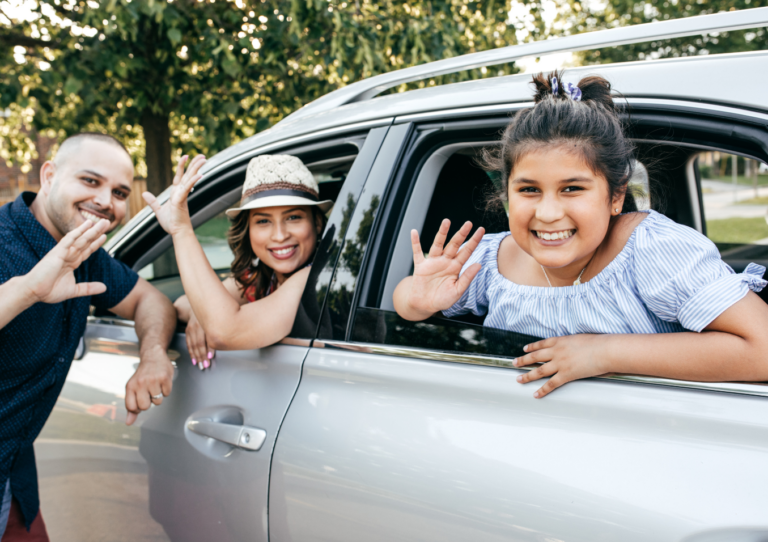 A male with close-cropped hair leaning against the driver's door of a silver vehicle while a woman with dark hair wearing a hat and a young girl with dark hair wave from the driver's and passenger's windows