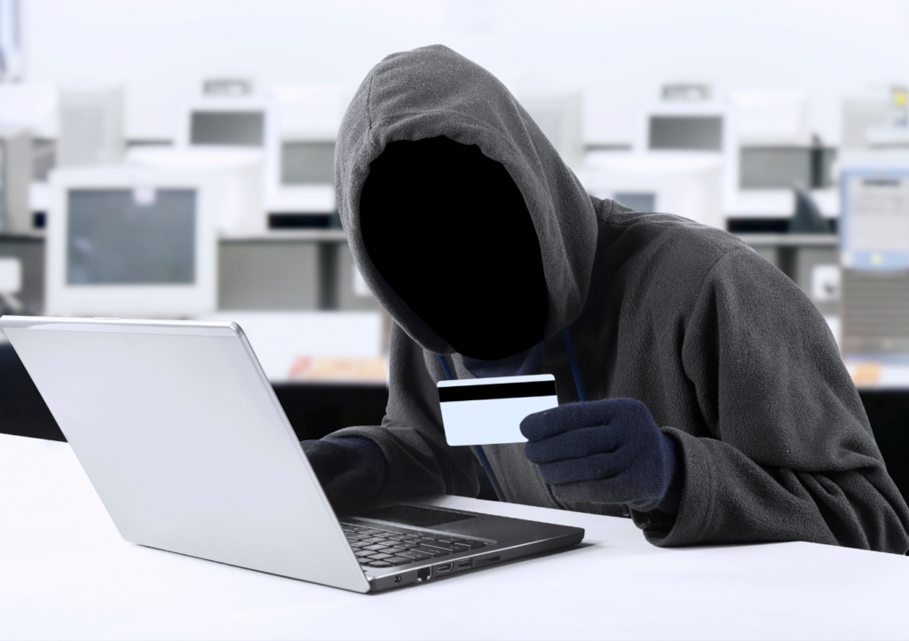 a faceless person wearing a hoodie and gloves, sitting in front of an open laptop and holding a credit card